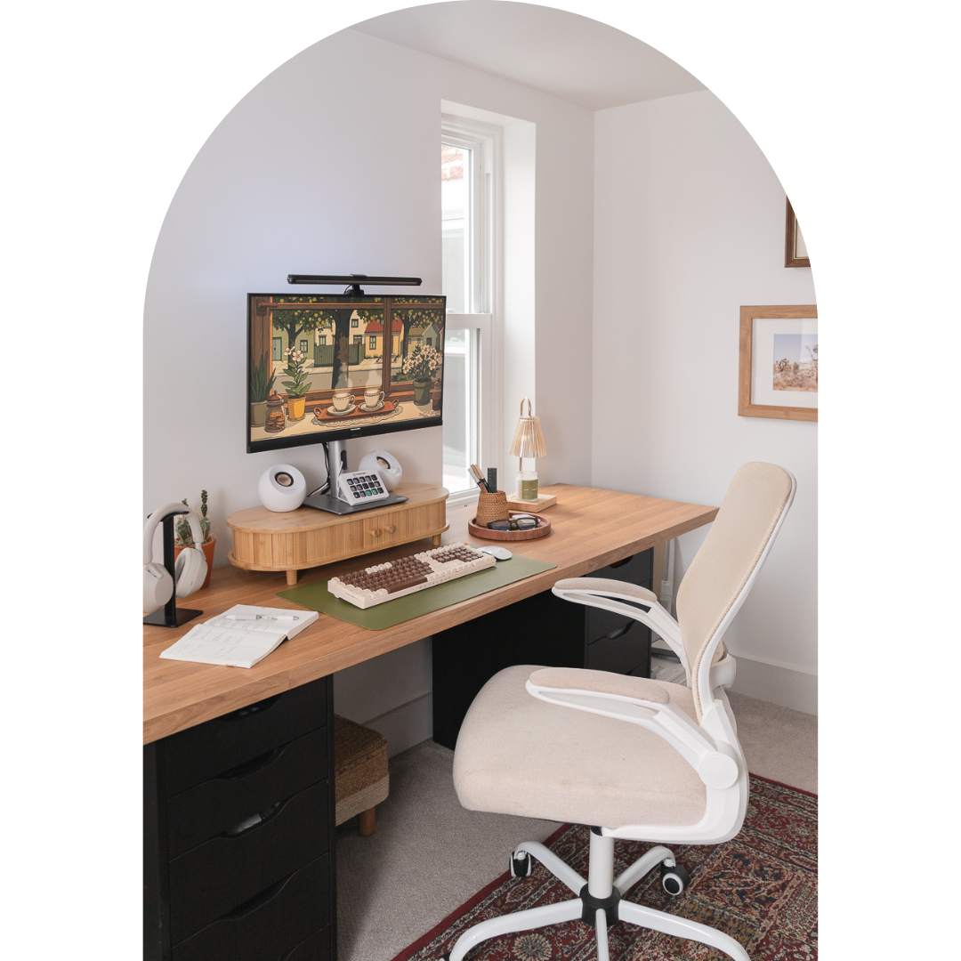 Boho-inspired home office setup with a wooden desk, ergonomic beige office chair, dual black drawer units, and a desktop monitor displaying digital art. Decor includes a green desk mat with a retro keyboard, white speakers, a rattan desk organizer, and framed botanical prints on the white wall.