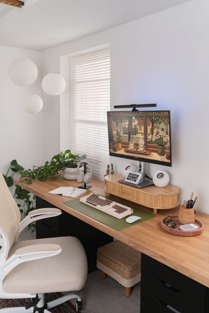 Boho-inspired home office setup with a wooden desk, ergonomic beige office chair, dual black drawer units, and a desktop monitor displaying digital art. Decor includes a green desk mat with a retro keyboard, white speakers, a rattan desk organizer, and a quarterly wall calendar.