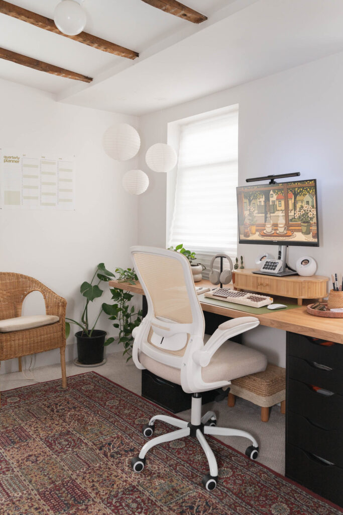 Boho-inspired home office setup with a wooden desk, ergonomic beige office chair, dual black drawer units, and a desktop monitor displaying digital art. Decor includes a green desk mat with a retro keyboard, white speakers, a rattan desk organizer, and a quarterly wall calendar.