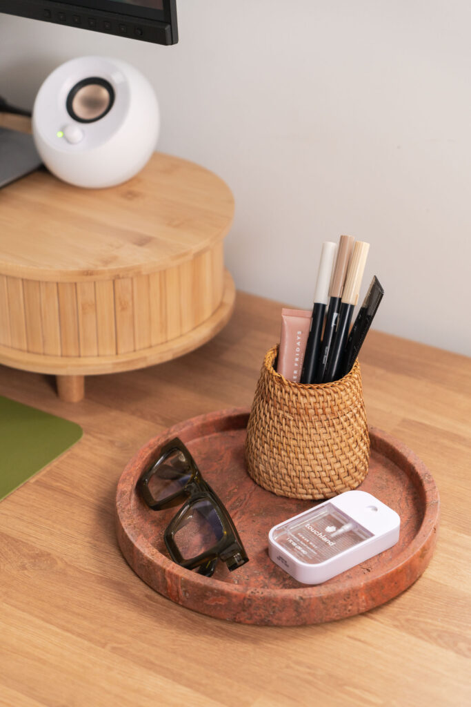 Close-up of a boho-style home office desk setup featuring a small woven pen holder with markers and lip balm, a red stone tray holding tortoiseshell glasses and a Touchland hand sanitizer, next to a bamboo monitor riser and white speaker.