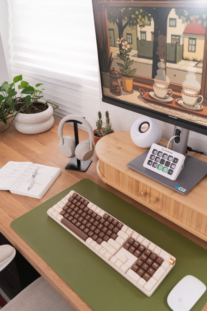 Close-up of a cozy home office desk setup with a brown and cream mechanical keyboard on a green desk mat, a white wireless mouse, a bamboo monitor riser holding a Stream Deck and speaker, and a monitor displaying cozy illustrated artwork. Also shown are neutral over-ear headphones on a stand, a white ceramic planter with a trailing plant, a small cactus, and an open weekly planner with a pen.