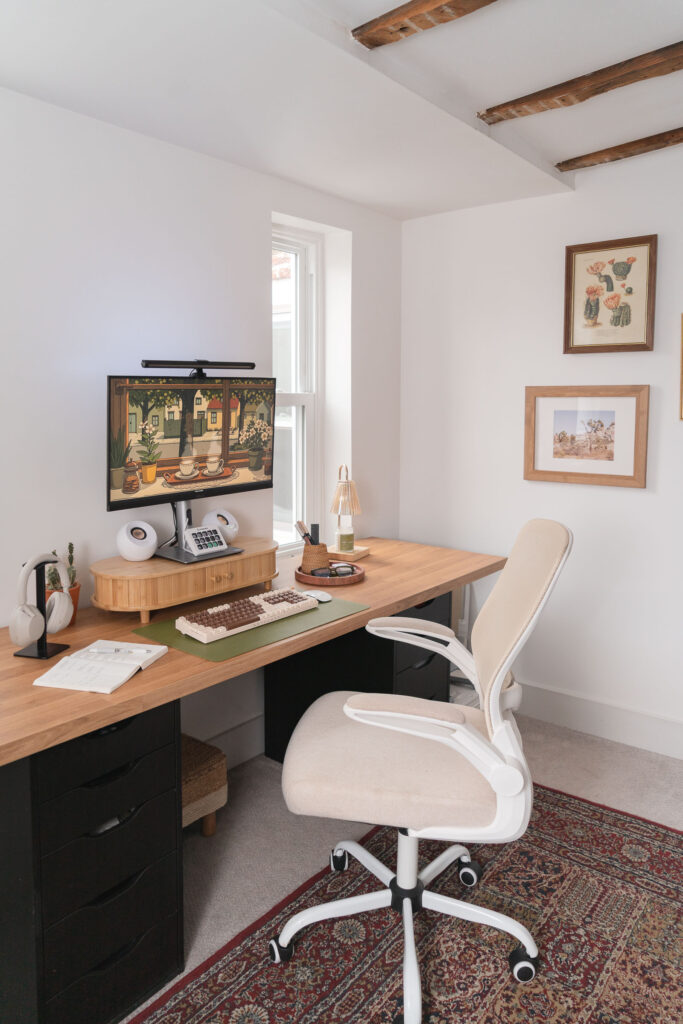 Boho-inspired home office setup with a wooden desk, ergonomic beige office chair, dual black drawer units, and a desktop monitor displaying digital art. Decor includes a green desk mat with a retro keyboard, white speakers, a rattan desk organizer, and framed botanical prints on the white wall.
