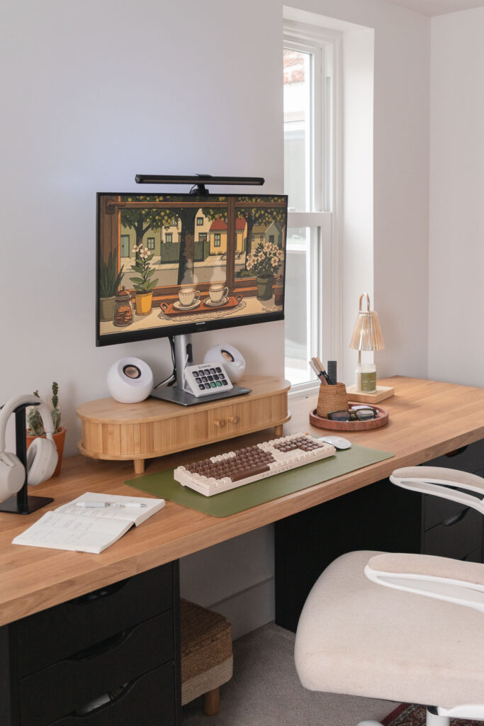 Boho-inspired home office setup with a wooden desk, ergonomic beige office chair, dual black drawer units, and a desktop monitor displaying digital art. Decor includes a green desk mat with a retro keyboard, white speakers, a rattan desk organizer, and framed botanical prints on the white wall.